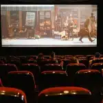 Sala cinematografica vuota con file di poltrone rivolte verso lo schermo illuminato in penombra.