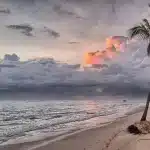 Libri colorati impilati su un asciugamano da spiaggia al sole, con occhiali da sole e mare sullo sfondo