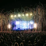 Palco illuminato di sera in piazza con folla in controluce e luci estive